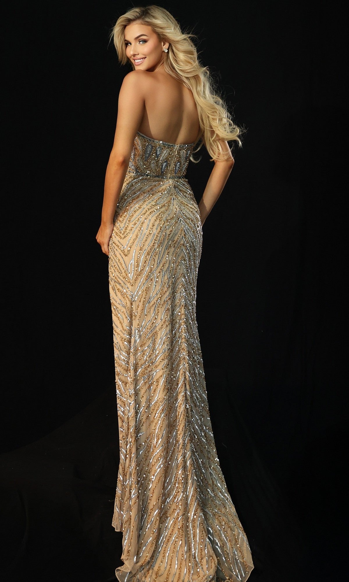 A woman with long blonde hair models the Jovani 46928, a fully embellished long prom dress with an asymmetrical sheer panel and high leg slit, smiling over her shoulder against a black background.